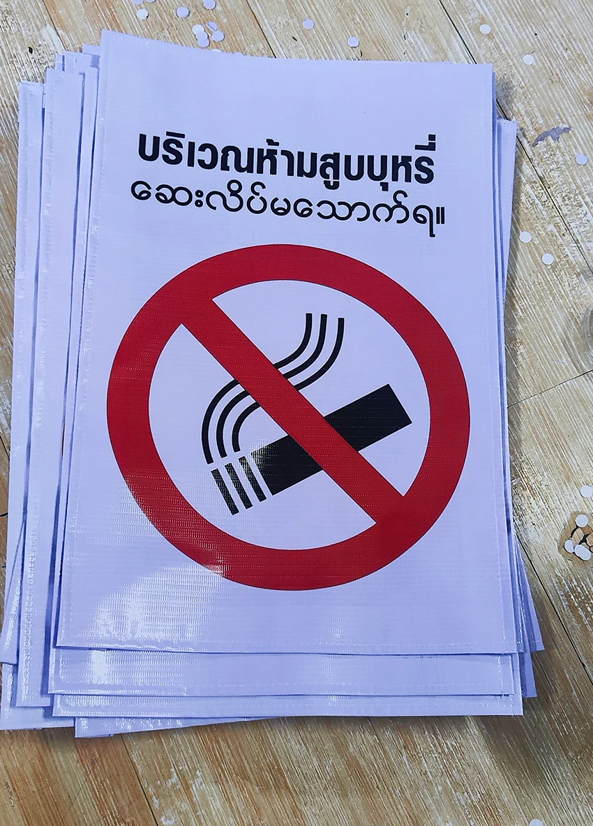 ป้ายไวนิลบริเวณห้ามสูบบุหรี่ พื้นขาว วงกลมแดงคาดสัญลักษณ์บุหรี่ ข้อความไทย–พม่า