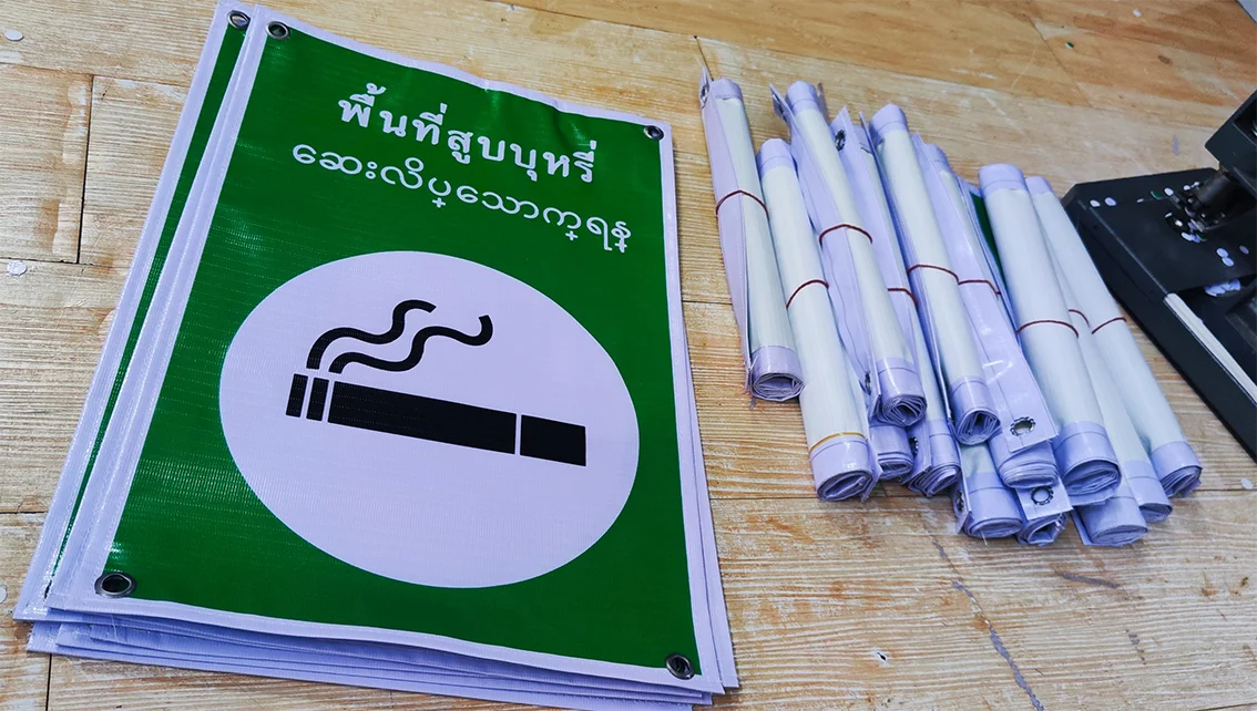 ป้ายไวนิลพื้นที่สูบบุหรี่ พื้นสีเขียว ตัวอักษรสองภาษา ไทย–พม่า พร้อมไอคอนบุหรี่